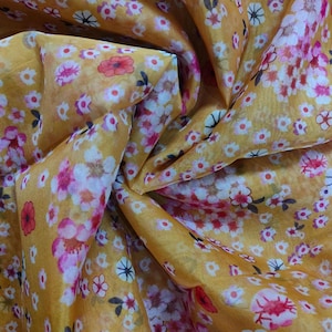 Puede incluir: Una tela transparente de color amarillo dorado con un patrón denso y repetitivo de pequeñas flores multicolores en tonos rosa, blanco y rojo oscuro. El material está recogido en pliegues, creando una superficie texturizada con luces y sombras.