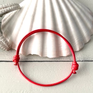 May include: A red string bracelet with a simple knot closure. The bracelet is photographed on a white seashell background.