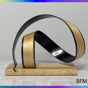 Op de afbeelding: Een houten sculptuur met een zwarte en natuurlijke houtkleurige afwerking. De sculptuur is gemaakt van twee stukken hout die met elkaar zijn verweven. De sculptuur staat op een houten voet.