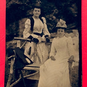 May include: Two women in vintage clothing pose outdoors. The woman on the left wears a long dress with a dark vest and a hat. The woman on the right wears a long white dress with a hat and sits on a wooden bench.