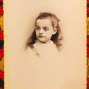 May include: A black and white portrait photograph of a young girl with long hair, wearing a white dress and a ribbon in her hair. The photo is framed with a decorative border.