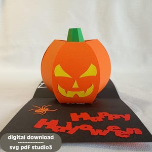 May include: A Halloween-themed pop-up card. The card features an orange pumpkin with a carved face and a green stem. The black card has the words "Happy Halloween" in red and a small orange spider.