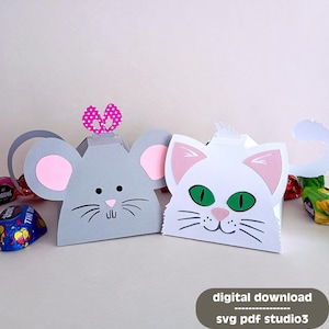Puede incluir: Dos cajas de manualidades de papel con forma de ratón gris y gato blanco. El ratón tiene orejas rosas y un lazo rosa. El gato tiene ojos verdes y orejas rosas. Las cajas están diseñadas para guardar pequeños dulces o regalos.