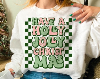 May include: White sweatshirt with a retro-style graphic that reads "Have a Holy Jolly Christmas" in pink and green bubble letters. The design includes a green and white checkered pattern. The sweatshirt is a festive holiday apparel item.