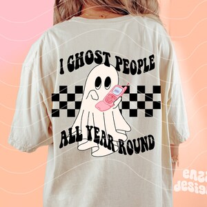 May include: Cream-colored t-shirt with a graphic of a ghost holding a pink flip phone. The text "I Ghost People All Year Round" is above and below the ghost, with a black and white checkered pattern.