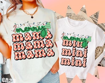 Op de afbeelding: Twee bijpassende crèmekleurige en witte T-shirts. Het eerste shirt heeft de woorden "merry mama mama mama" in een retro lettertype met een kerstthema. Het tweede shirt heeft de woorden "merry mini mini mini" in een vergelijkbare stijl.