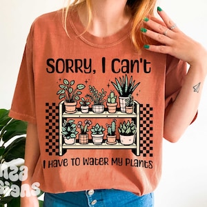 Puede incluir: Una camiseta de color &oacute;xido con el texto "Lo siento, no puedo, tengo que regar mis plantas". El texto est&aacute; en negro y est&aacute; sobre un dibujo de una estanter&iacute;a con plantas en macetas.