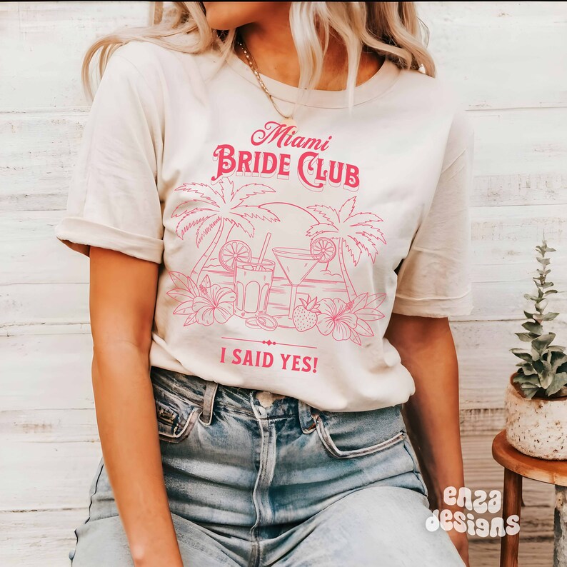 May include: A white t-shirt with a pink graphic design that says "Miami Bride Club" with palm trees, cocktails, and the text "I said yes!"