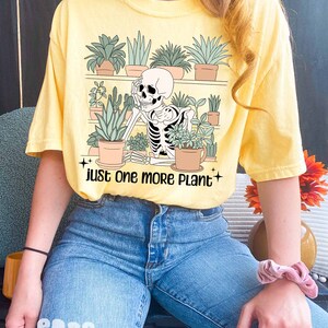 May include: A yellow t-shirt with a skeleton surrounded by potted plants. The text on the shirt reads "Just one more plant".