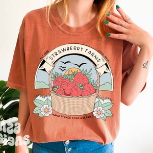 以下が含まれることがあります： さび色のTシャツに、イチゴの入ったバスケットのグラフィックデザインが施されています。デザインには、「Strawberry Farms」というテキストと、その両側に「Est. 1989」と「Local Fresh」という単語が書かれています。「Hand Picked Strawberries」というテキストは、イチゴの入ったバスケットの下にあります。