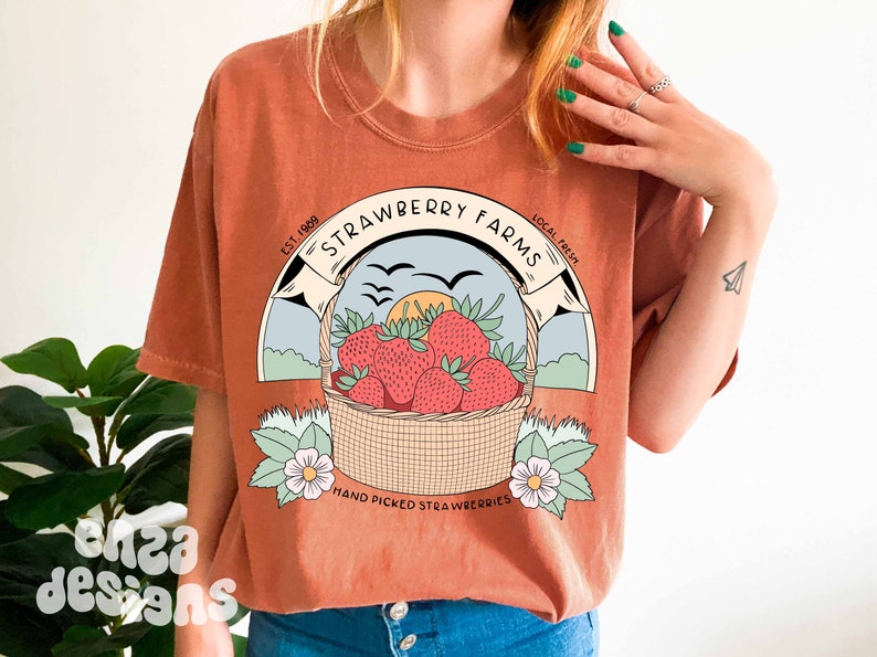 以下が含まれることがあります： さび色のTシャツに、イチゴの入ったバスケットのグラフィックデザインが施されています。デザインには、「Strawberry Farms」というテキストと、その両側に「Est. 1989」と「Local Fresh」という単語が書かれています。「Hand Picked Strawberries」というテキストは、イチゴの入ったバスケットの下にあります。
