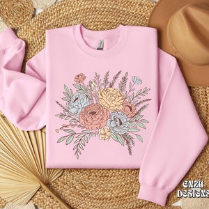 May include: A pink sweatshirt with a floral design featuring a bouquet of pastel-colored flowers and green leaves. The text "ENZA DESIGNS" is printed on the bottom right corner of the sweatshirt.
