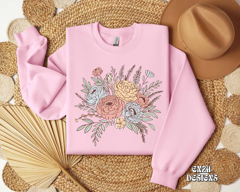 May include: A pink sweatshirt with a floral design featuring a bouquet of pastel-colored flowers and green leaves. The text "ENZA DESIGNS" is printed on the bottom right corner of the sweatshirt.