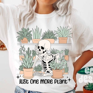 May include: White t-shirt with a graphic of a skeleton sitting among potted plants on a shelf. The text "just one more plant" is printed below the graphic.
