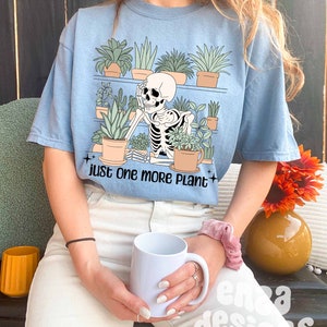 May include: A light blue t-shirt with a graphic of a skeleton surrounded by potted plants. The text "just one more plant" is printed below the graphic.