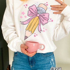 May include: White sweatshirt with a yellow pencil tied with a pink bow and surrounded by pink and purple stars. The text "ENZA DESIGNS" is printed on the bottom of the sweatshirt.