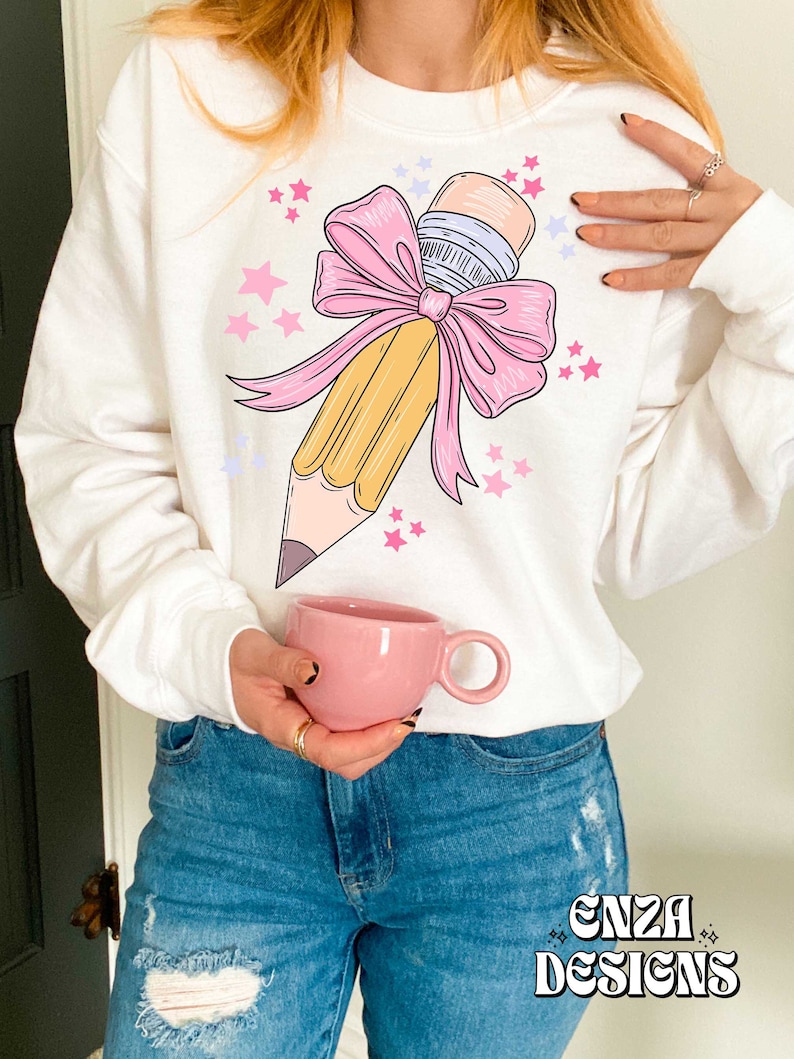 May include: White sweatshirt with a yellow pencil tied with a pink bow and surrounded by pink and purple stars. The text "ENZA DESIGNS" is printed on the bottom of the sweatshirt.