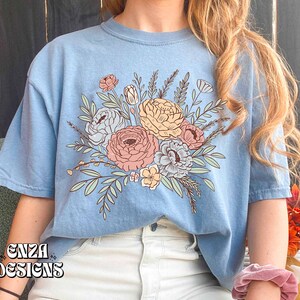 May include: A light blue t-shirt with a floral design featuring pink, yellow, and white flowers with green leaves. The design is centered on the front of the shirt.