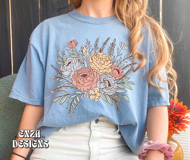 May include: A light blue t-shirt with a floral design featuring pink, yellow, and white flowers with green leaves. The design is centered on the front of the shirt.