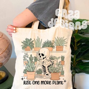 May include: A white tote bag with a skeleton surrounded by potted plants. The text "just one more plant" is printed on the bag.