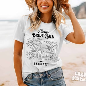 May include: White t-shirt with a black graphic design that says "Miami Bride Club" with palm trees and cocktails. The text "I said yes!" is below the graphic.