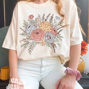 May include: A cream colored t-shirt with a floral design featuring pink, blue, and yellow flowers with green leaves.