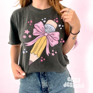 May include: A dark gray t-shirt with a graphic of a yellow pencil tied with a pink bow and surrounded by pink stars.