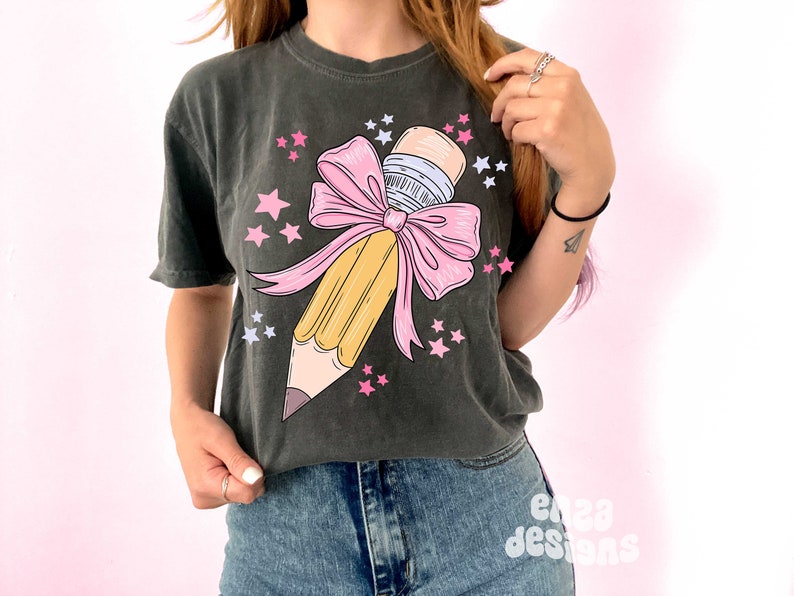 May include: A dark gray t-shirt with a graphic of a yellow pencil tied with a pink bow and surrounded by pink stars.
