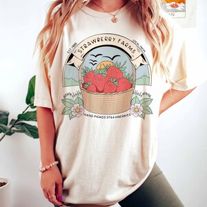 以下が含まれることがあります： クリーム色のTシャツを着た女性。ヴィンテージスタイルのイチゴのバスケットのグラフィックと「Strawberry Farms Est. 1989 Local Fresh Hand Picked Strawberries」のテキストがプリントされています。彼女は、脚に「enza designs」のテキストが書かれたダークグリーンのサイクリングショーツも着用しています。