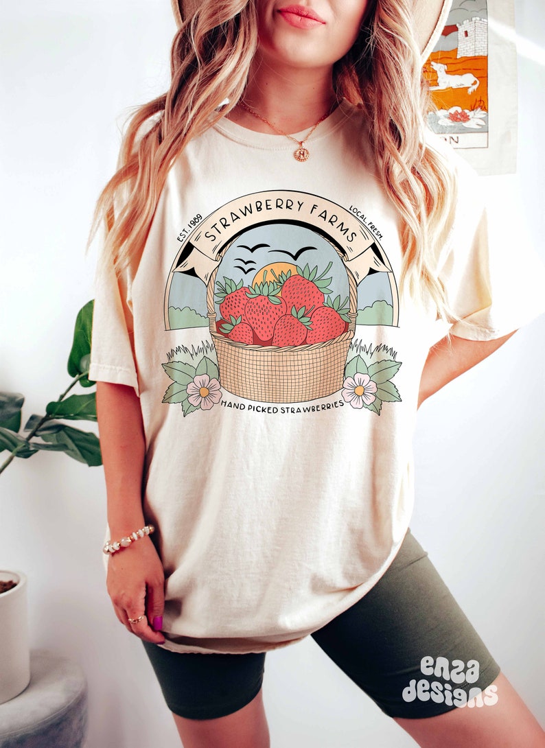 以下が含まれることがあります： クリーム色のTシャツを着た女性。ヴィンテージスタイルのイチゴのバスケットのグラフィックと「Strawberry Farms Est. 1989 Local Fresh Hand Picked Strawberries」のテキストがプリントされています。彼女は、脚に「enza designs」のテキストが書かれたダークグリーンのサイクリングショーツも着用しています。