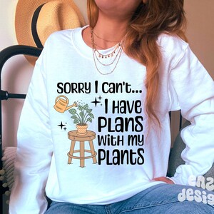 May include: White long-sleeve sweatshirt with the text "Sorry I can't... I have plans with my plants."  A cartoon illustration of a plant on a stool with a watering can pouring water on the plant.