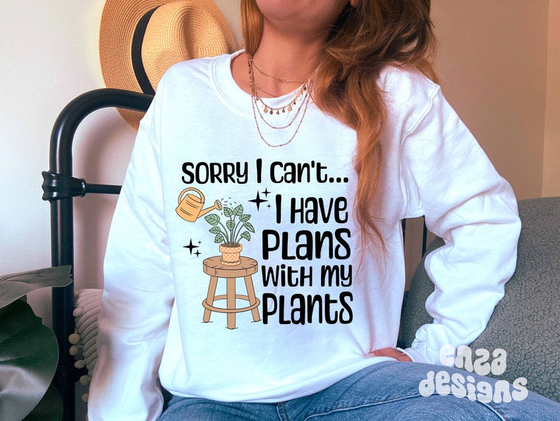 May include: White long-sleeve sweatshirt with the text "Sorry I can't... I have plans with my plants."  A cartoon illustration of a plant on a stool with a watering can pouring water on the plant.