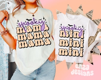 Può includere: Due magliette, una color crema e una bianca, a tema Halloween. La maglietta color crema dice "spooky mama mama mama" in lettere viola e arancioni. La maglietta bianca dice "spooky mini mini mini" in colori simili. Entrambe le magliette hanno grafiche di pipistrelli e castelli.