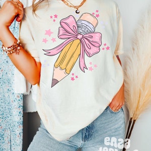 May include: A light yellow t-shirt with a graphic of a yellow pencil tied with a pink bow and surrounded by pink stars.