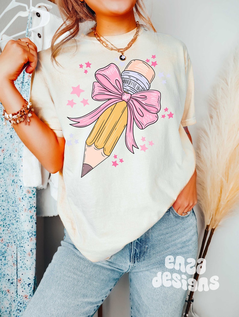May include: A light yellow t-shirt with a graphic of a yellow pencil tied with a pink bow and surrounded by pink stars.
