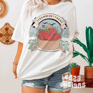 以下が含まれることがあります： イチゴの入ったバスケットのグラフィックデザインが施された白いTシャツ。デザインには「Strawberry Farms」というテキストと、テキストの両側に「Est. 1989」と「Local Fresh」という単語が含まれています。デザインには、「Hand Picked Strawberries」というテキストも下部に含まれています。