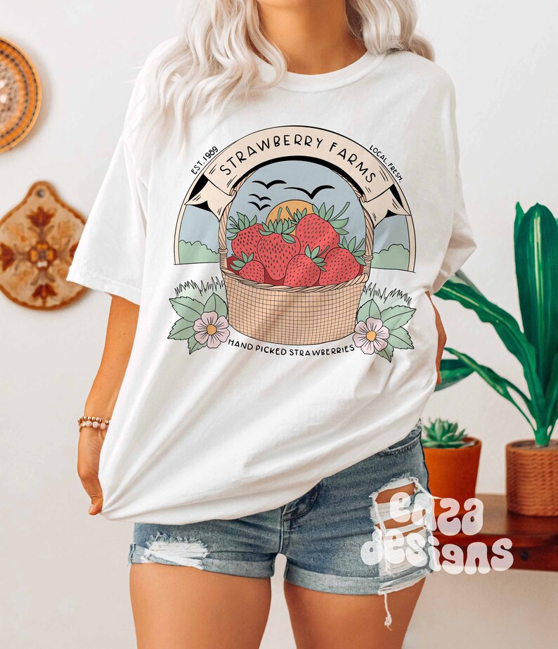 以下が含まれることがあります： イチゴの入ったバスケットのグラフィックデザインが施された白いTシャツ。デザインには「Strawberry Farms」というテキストと、テキストの両側に「Est. 1989」と「Local Fresh」という単語が含まれています。デザインには、「Hand Picked Strawberries」というテキストも下部に含まれています。