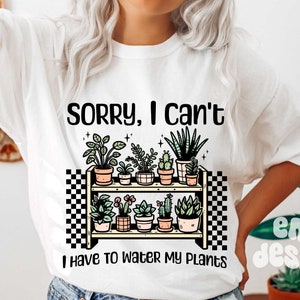 Puede incluir: Camiseta blanca con el texto "Lo siento, no puedo, tengo que regar mis plantas". El texto est&aacute; en negro y hay un gr&aacute;fico de un estante con plantas en macetas.