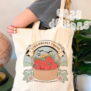 以下が含まれることがあります： 赤いイチゴ、夕日、鳥が描かれた茶色の円の中に「Strawberry Farms」と書かれたロゴが入った白いキャンバス地のトートバッグ。円のすぐ下には「Est. 1989 Local Fresh Hand Picked Strawberries」と書いてあります。