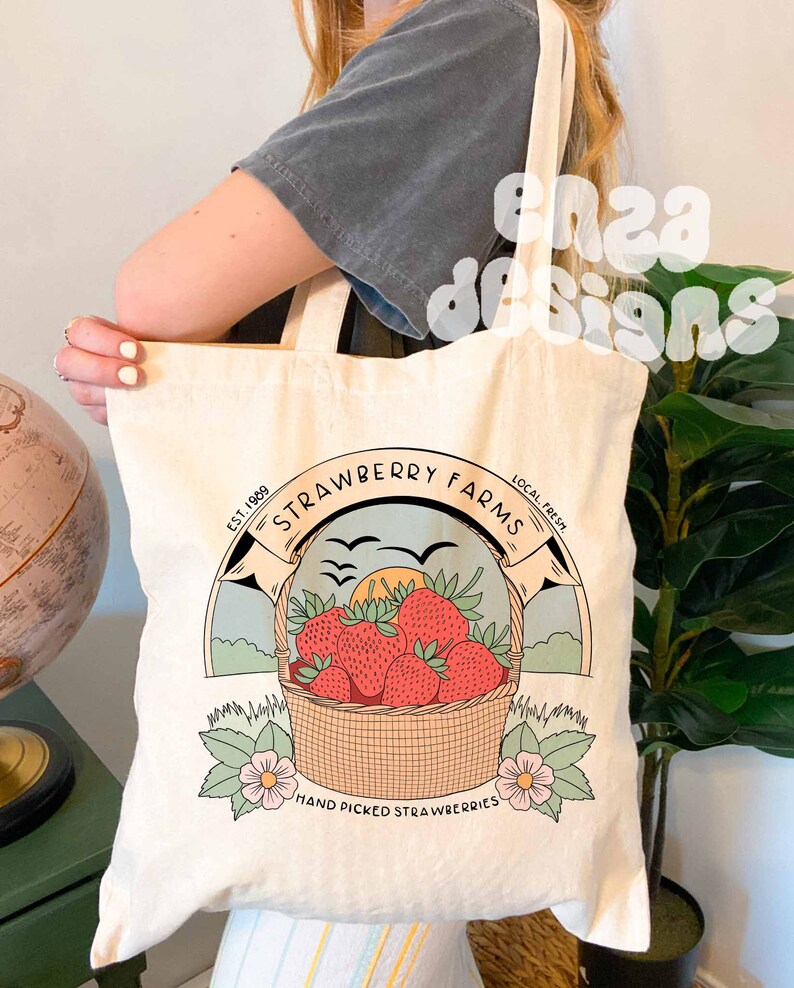 以下が含まれることがあります： 赤いイチゴ、夕日、鳥が描かれた茶色の円の中に「Strawberry Farms」と書かれたロゴが入った白いキャンバス地のトートバッグ。円のすぐ下には「Est. 1989 Local Fresh Hand Picked Strawberries」と書いてあります。