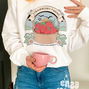 以下が含まれることがあります： 「Strawberry Farms」と書いてあるヴィンテージ風のロゴがプリントされた白いスウェットシャツ。赤いイチゴの入ったバスケット、飛んでいる鳥、そして「Est. 1989 Local Fresh Hand Picked Strawberries」という文字が書かれている。