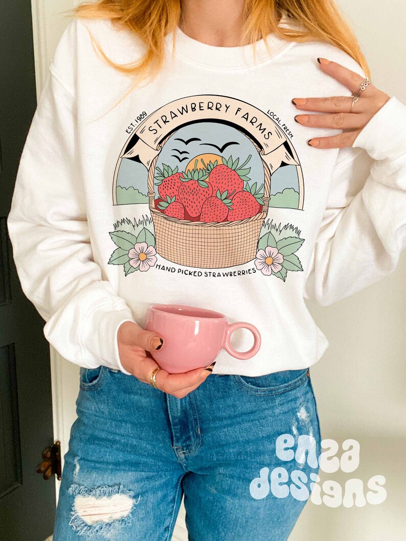 以下が含まれることがあります： 「Strawberry Farms」と書いてあるヴィンテージ風のロゴがプリントされた白いスウェットシャツ。赤いイチゴの入ったバスケット、飛んでいる鳥、そして「Est. 1989 Local Fresh Hand Picked Strawberries」という文字が書かれている。