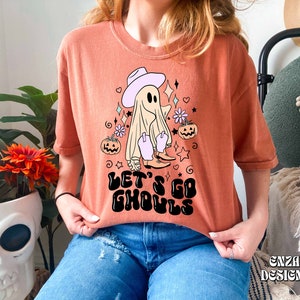 May include: Orange t-shirt with a Halloween-themed graphic. The design features a ghost wearing a cowboy hat and boots, surrounded by stars, flowers, and pumpkins. The text "LET'S GO GHOULS" is printed in black. The shirt is a relaxed fit.