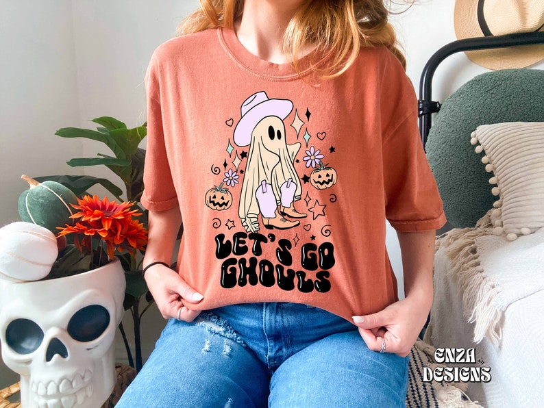 May include: Orange t-shirt with a Halloween-themed graphic. The design features a ghost wearing a cowboy hat and boots, surrounded by stars, flowers, and pumpkins. The text "LET'S GO GHOULS" is printed in black. The shirt is a relaxed fit.