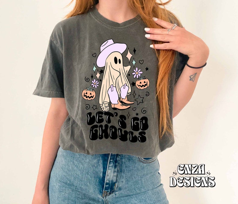 May include: Dark gray t-shirt with a Halloween-themed graphic. The design features a ghost wearing a cowboy hat and boots, with the text "LET'S GO GHOULS". The design includes pumpkins, stars, and flowers.