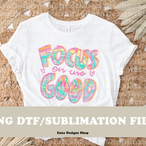 May include: White t-shirt with the text "Focus on the Good" in colorful, swirled lettering. The design includes hearts and is a PNG DTF/Sublimation file. The shirt is displayed on a woven background.