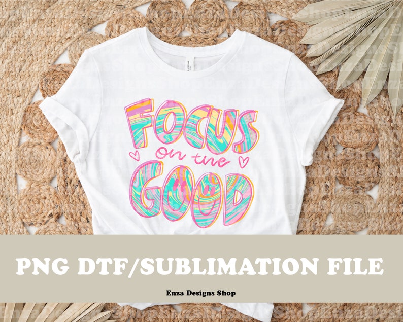 May include: White t-shirt with the text "Focus on the Good" in colorful, swirled lettering. The design includes hearts and is a PNG DTF/Sublimation file. The shirt is displayed on a woven background.