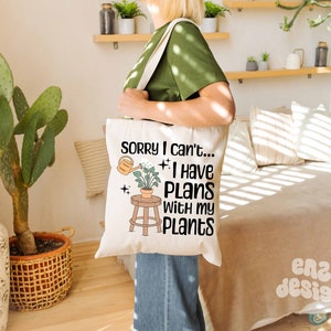 May include: A white tote bag with a black graphic that says "Sorry I can't... I have plans with my plants". The graphic also includes a small potted plant and a watering can.