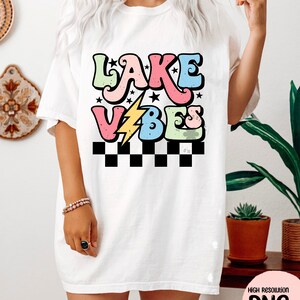 Puede incluir: Una camiseta blanca con un dise&ntilde;o gr&aacute;fico colorido que dice "Lake Vibes" en una fuente retro. El gr&aacute;fico incluye un rayo y estrellas.