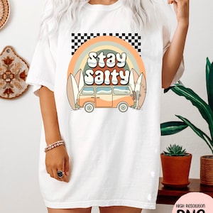 May include: A white t-shirt with a graphic design of a vintage van driving past surfboards with a rainbow and the text "Stay Salty" in a retro font.